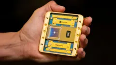 A hand holding the Majorana 2 chip in its palm. Including its housing the chip appears to be about the size of a beer mat. It consists of a gold plated metal frame and sits on a dark blue-green board bearing its name. The chip itself is a small silvery square on a dark gunmetal blue square with very fine gold circuitry visible.