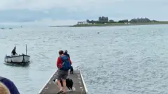 A pier stretching out to the sea with people and a border collie waiting for a boat. The boat is a large rowing boat to the left and a man is steering it. In the background, Piel Island can be seen - it has rocky ruins and a few small buildings.