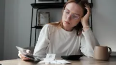 Getty Images A woman sitting at a table with her head in her hands, a cup of tea and a calculator in front of her, looking at receipts.