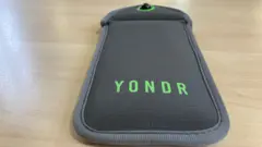 A brown pocket sleeve wallet rests on a wooden table. The company name Yondr is written on it in green colour. It is designed to store a mobile phone device with a magnetic seal that can be reopened at a later time or date
