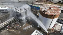 Water is sprayed on a stack of four cylindrical storage tanks, with another larger brown and white tank visible alongside them.