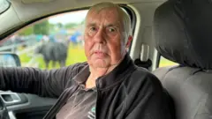 A man with white hair wearing a black polo and black jacket
