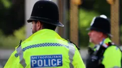 Two police officers in high viz face away from the camera and they have "Metropolitan Police" printed on their backs. One man has his back to the camera and another looks to the left. Both wear helmets.