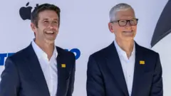 Apple's incoming chief executive John Ternus standing next to his predecessor Tim Cook. Both are smiling for cameras at an Apple event and wearing an almost identical blue blazer and white collared shirt.