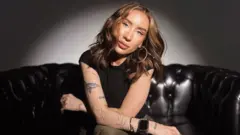 Katya has long mid-brown hair and blue eyes. She is wearing big hoop earrings and has her nose pierced. She has tattooes in the shape of plants and butterflies down one arm. 