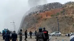Riot police stood at the bottom of a cliff, with dozens of migrants stood on it