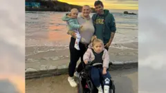 Charlie Brown Charlie Brown stands shoulder to shoulder with his family, including Dani who is in a wheelchair. He is posing in front of the camera smiling. They are standing on a sandy beach with the shore and shallow waves behind them. Charlie has a green hoodie and short brown hair, next to him is a woman wearing a cream hoodie and black leggings with white Crocs. She's wearing a mint green jacket and a white onesie with colorful dinosaurs on it, holding a baby in her arms.