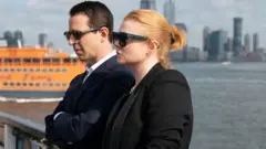 Warner Brothers Discovery Jeremy Strong and Sarah Snook from Succession stand by the water in sunglasses and suits, with New York City in the background