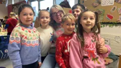 Gwen Rose smiling with five children. They are all wearing Christmas jumpers.
