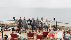 Eight performers stand in a line on the promenade and sing into microphones. There is a crowd of people on the stairs and in deck chairs watching on. the sea behind them is grey and calm, and a similar colour to the sky above.