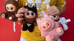 A boy in a yellow sports t-shirt holds up four Labubu dolls. The furry gremlin-like dolls have big glossy eyes and toothy grins. You can't see Harri's head but his chest and the focus is on the dolls. There is a a brown one on the far left, another brown one with black Prada dungarees on, a pink one next to that and then a white one holding a Coca Cola can on the far right.