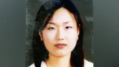 Jong-Ok Shin in what appears to be a copy of a family photo, where the folds and marks on the print are still visible, showing a close-up of a young women with long, dark hair and small hooped earrings looking straight at the camera with a neutral expression.