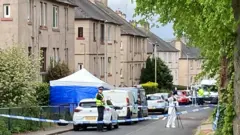A cordoned off area of a street in South Queensferry. There are people wearing white hazmat suits and uniformed police officers. A blue forensic tent is in the front garden of one of the properties and cars line the street.
