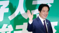 Taiwanese President Lai Ching-te stands to the right of the picture waving and smiling at his party HQ in Taipei. Behind him is a green backdrop with Chinese lettering.