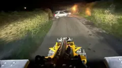 A view from what appears to be a combine harvester or some other farm machinery, driving along a narrow road seemingly lit only by the headlights, with the hedgerows either side blurred slightly from the movement. Ahead on the road is what appears to be a silver SUV, which by its position appears to have emerged from a gate to the left of the image.