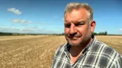 David Lemon, a man with greying hair and stubble, wearing a checked shirt, stands in one of the large fields of his farm against a blue sky with a few small clouds.