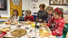 Iris and four children putting butter and jam on slices of bread. They are all working around the same table and wearing Christmas jumpers.