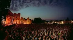 A large crowd of people in the dark with a castle illuminated in the background and a stage on the left hand side