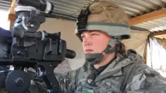 Nadine Askew wearing a camouflage military uniform and rounded helmet with a chin strap. She is sitting underneath a corrugated metal shelter, looking towards the left as she aims a machine gun.