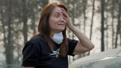 A woman holds her head with her hand. Her hands are closed and she has a face mask loose around her neck.