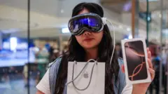 A woman standing in what looks like a bright, modern tech store. Theyu2019re wearing a large reflective mixedu2011reality headset with a cable trailing from it, the kind that covers the upper half of the face like a visor. In one hand theyu2019re holding an Apple shopping bag, and in the other theyu2019re carrying a boxed iPhone. The background is softly blurred, with glass panels and indistinct figures suggesting a busy retail environment
