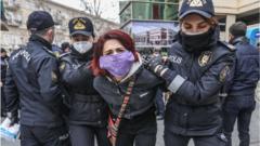 Две женщины полицейские ведут героиню истории, скрутив ей руки за спиной. На ней медицинская маска с надписью: "Сражайся как женщина"
