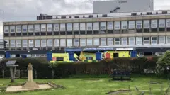 David Robinson/Geograph In the center stands a long, wide building with many windows, with ambulances outside the entrance, a sign reading 'Darlington Memorial Hospital'. There is a park in front.