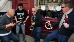 Image of four band members from the group Madness, wearing jeans and cardigans and performing on the street outside a pub. One band member is playing a ukulele, a second plays a saxophone and the lead singer Suggs has a microphone and tambourine. The other has no instrument but is standing, smiling.