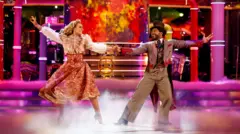 The dance partners were dressed in fancy Victorian style costumes, dancing in the ballroom with pink lights and smoke all around. JB is wearing a brown suit with a waistcoat and top hat, while Amy is wearing a dress with a white blouse on top and an embroidered peach-colored skirt.