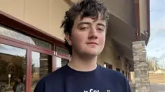 Darian Quist in a navy blue T-shirt looks at the camera with a neutral expression. Behind him is a community centre.