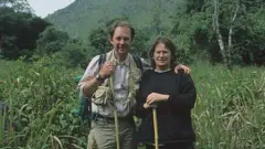 Bleddyn and Sue Wynn Jones in tall grass with trees and mountains in the background