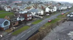 A aerial shot of the seafront road at Beadnell. Large houses are on one side of the road, facing a pebbly beach on the other. Behind are a few hundred other houses with the sea and a stretch of the coastline in the top far right.