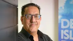 Nazir Afzal, who wears dark glasses and a black shirt, smiles as he leans against a dark wall in front of a blue banner for BBC's Desert Island Discs.
