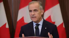 An image of PM Mark Carney wearing a suit, speaking at a podium. Behind him are three poles holding Canadian flags. He is photographed mid-speech. He has short black and grey hair. 