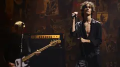 Getty Images The sombre half-open black shirt and black leather trousers hold a microphone stand and look moody on stage next to a guitarist at the 2025 MTV Video Music Awards.