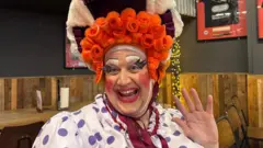 Dion Davies wears his Lucie Goose costume, an orange wig, with a spotty top and a bonnet and ribbon.
