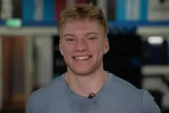 Close-up of a person in a light blue sports top, facing the camera and smiling. The background is softly blurred, showing an indoor gym. 