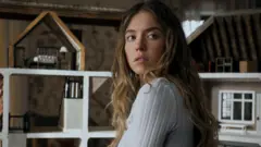 Lionsgate Sydney Sweeney appears with her head turned to face away from the camera. She is wearing a white long-sleeved ribbed top and is in front of a dolls house.