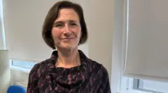 Annette Morris has short, brown hair wearing a dark purple and black patterned dress with a cowl neck in an office setting. 