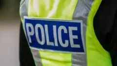 A police officer shoulders and back pictured from behind. They are wearing a black top and a yellow hi-vis gilet with the word police in white on a blue background.