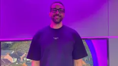 A man with glasses and a blue shirt stands in a studio and smiles while bathed in purple light.