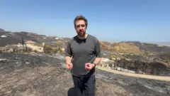 Nick Beake in a black shirt standing in the aftermath of wildfire.