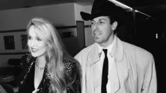 Getty Images A black and white photograph of model Jerry Hall wearing a shiny jacket and black hat and trench coat with fashion designer Antony Price in London in 1985.