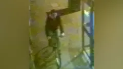 A grainy still from CCTV which shows a man wearing a dark jacket cycling on the pavement past a phone box