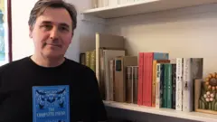 Alma Books Alessandro Galenzi, a man in a black sweater with short brown hair, holding a book of poetry by Dylan Thomas next to a bookshelf