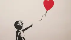 Banksy print of Girl with Balloon is on the right of the picture and a blue phone light is on the left, where someone is taking a photo of it. The background is black. Girl with Balloon is in a white frame. It shows a spray stencil style image of a young girl in a dress reaching for the string of a balloon as it soars away. The balloon is a red love heart.