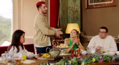Tom standing at the Traitors castle's breakfast table and gesturing at Alex, who's seated next to him