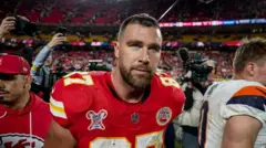Bearded Travis Kelce is seen in the centre of the image wearing Kansas City Chiefs' red home jersey as he leaves the field in a sports stadium. Behind him TV camera operators are trying to film him.