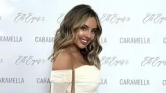 Caramella founder Mollie-May Rossi smiles in front of a branded white backdrop sporting her company name. She is wearing a shoulderless white dress with sleeves and has a grey handbag with gold handles over one shoulder.