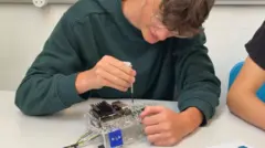 A teenage boy in a gark green jumper with short brown hair and glassed works witha screwdriver on a small platic and metal appliance.